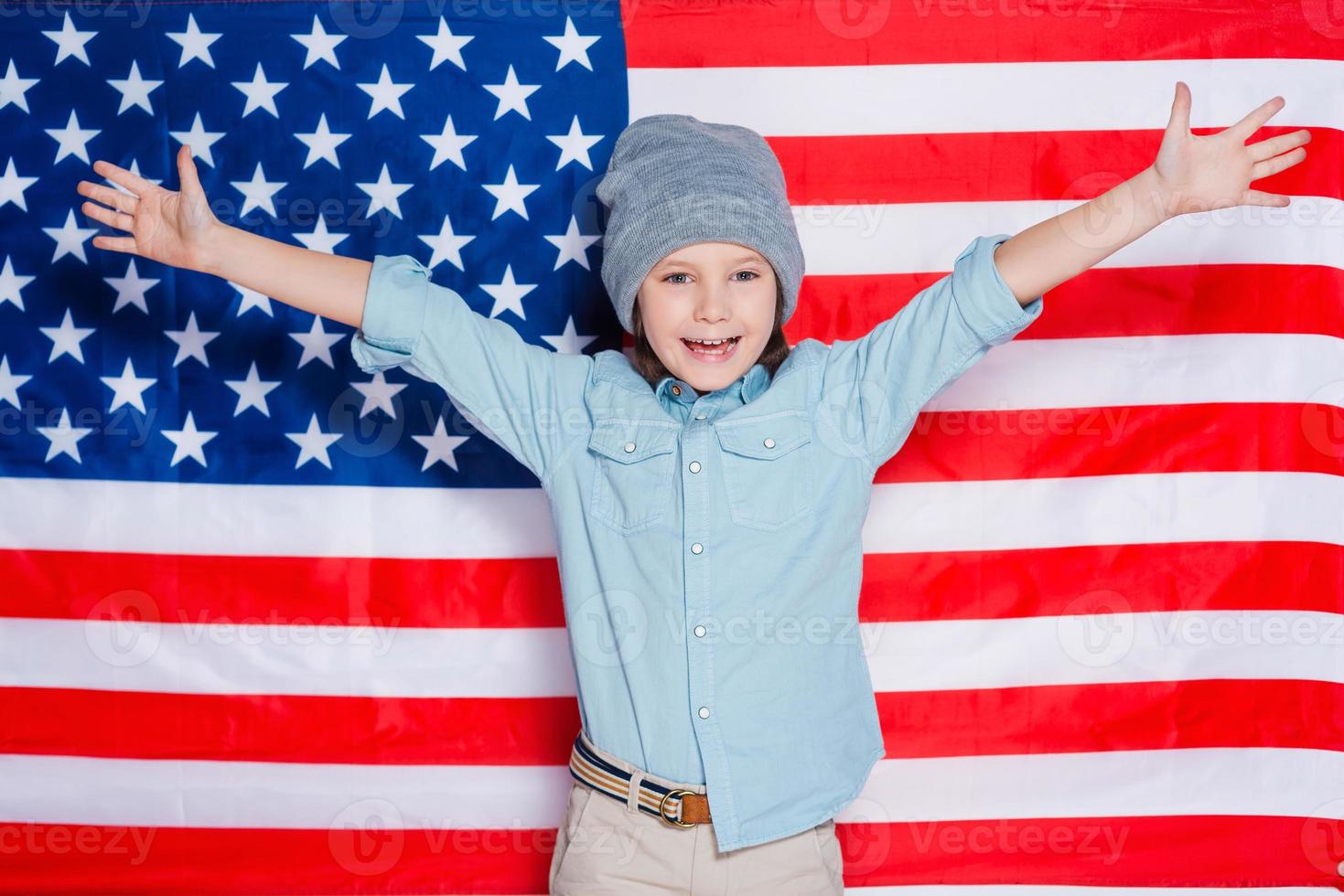 bonjour amérique petit garçon en lunettes gardant les bras levés et souriant debout contre le drapeau américain photo