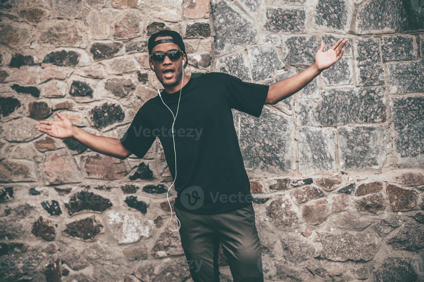 quel beau jeune homme africain dans les écouteurs regardant la caméra et faisant des gestes tout en se tenant devant le mur de pierre à l'extérieur photo