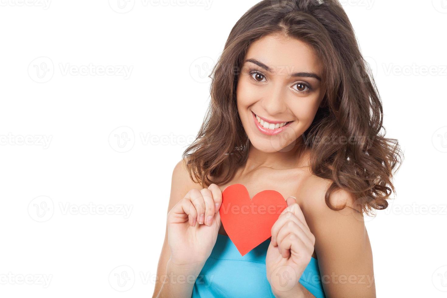 je suis amoureux. Jolie jeune femme tenant un coeur de papier rouge et souriant à la caméra en se tenant debout isolé sur fond blanc photo