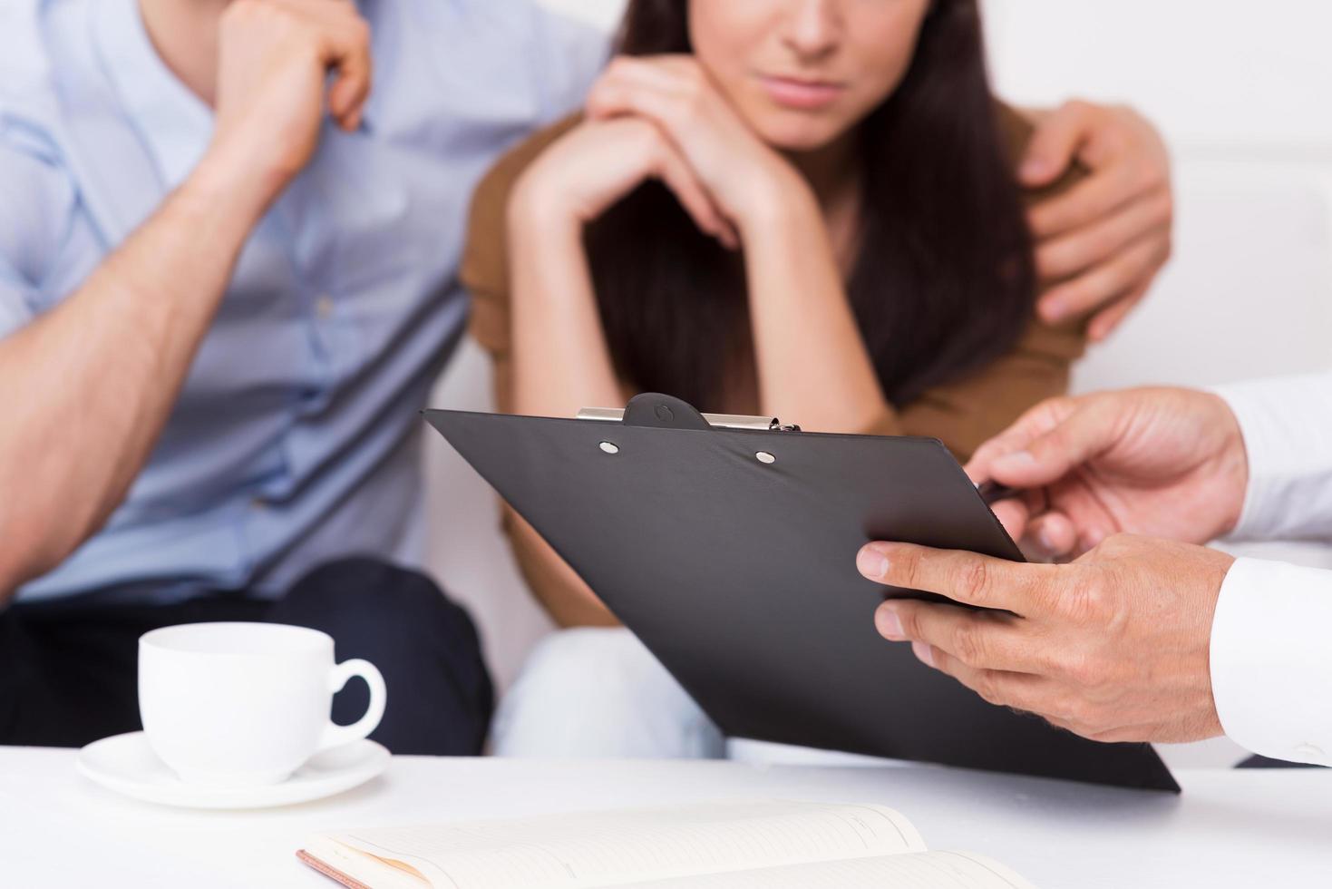 signez ici. gros plan d'un jeune couple réfléchi assis sur le canapé tandis que l'homme en tenue de soirée pointant le presse-papiers avec le document photo