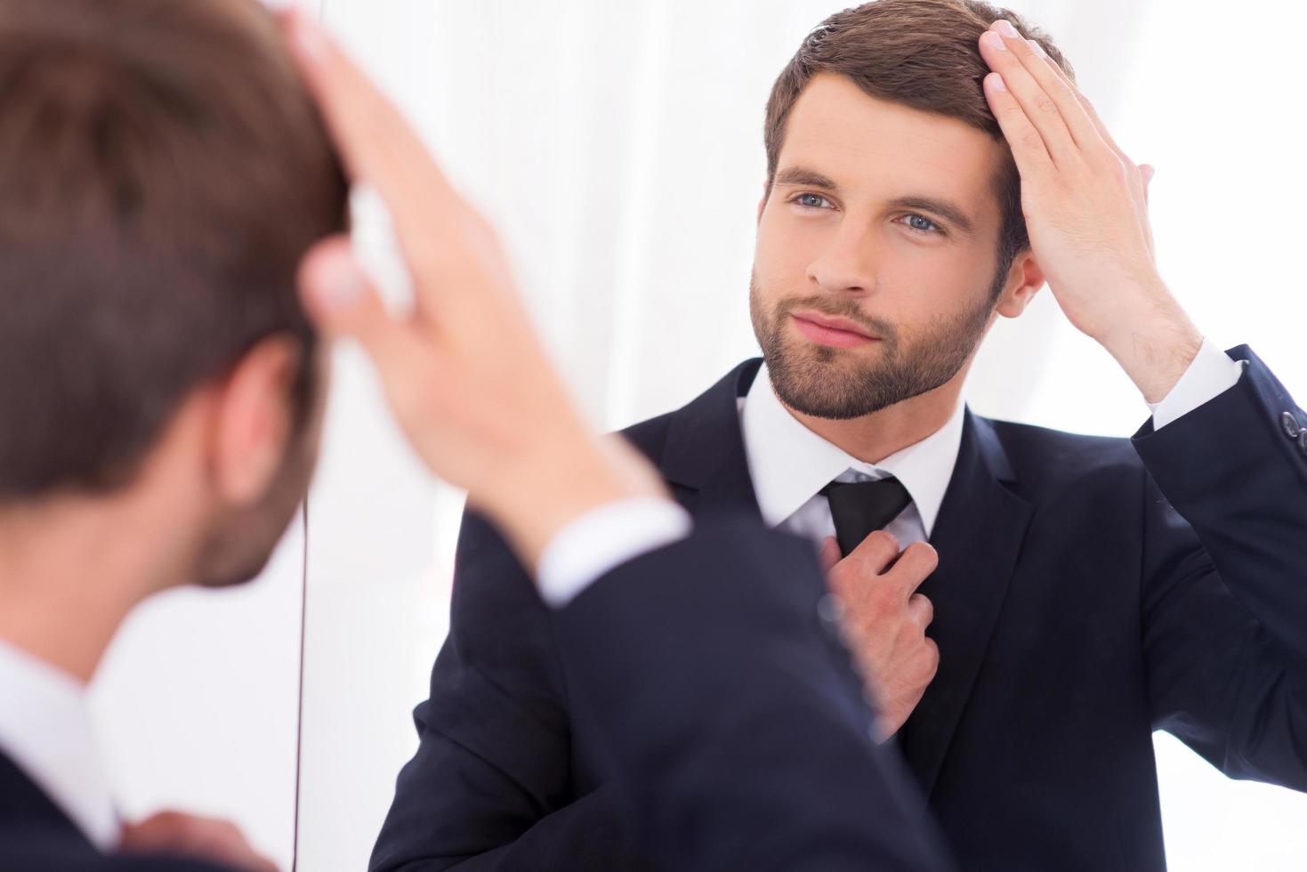 en s'assurant qu'il a l'air parfait. beau jeune homme en tenues de soirée ajustant sa coiffure et souriant tout en se tenant contre le miroir photo