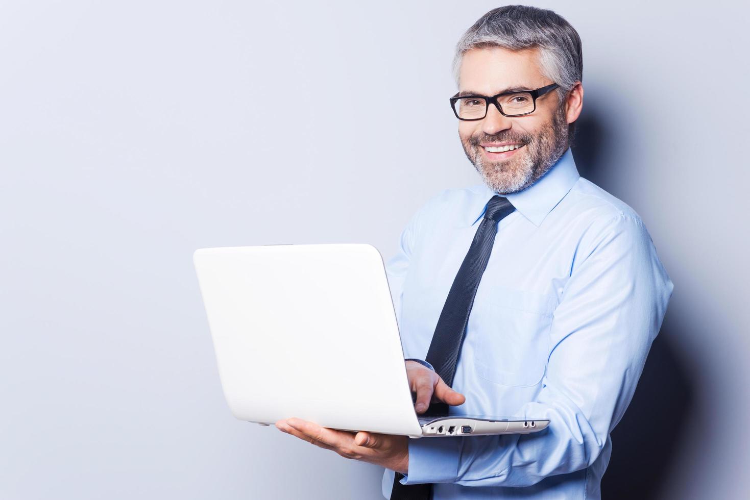 soutenir votre entreprise. Confiant homme mûr en tenues de soirée travaillant sur ordinateur portable en se tenant debout isolé sur fond blanc photo