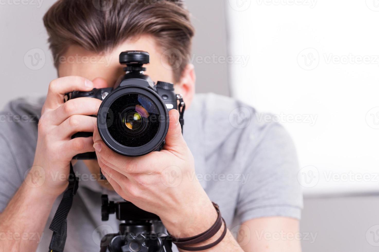 sourire jeune homme se concentrant sur vous avec un appareil photo numérique tout en se tenant en studio avec un équipement d'éclairage sur fond