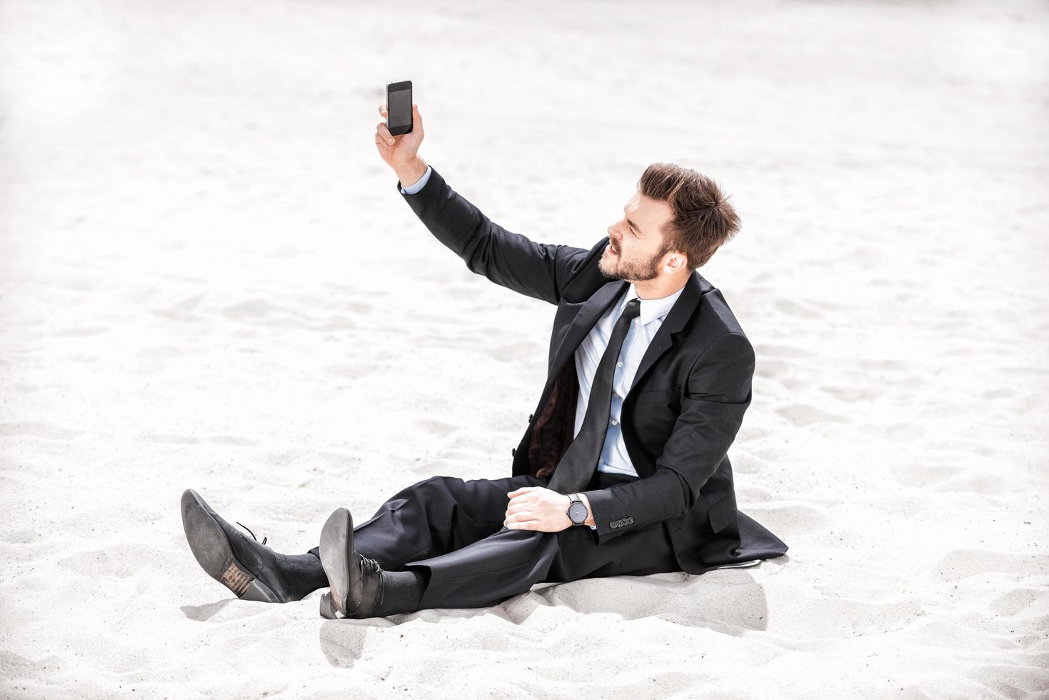 signal faible. jeune homme d'affaires frustré à la recherche d'un signal de téléphone portable assis sur le sable dans le désert photo