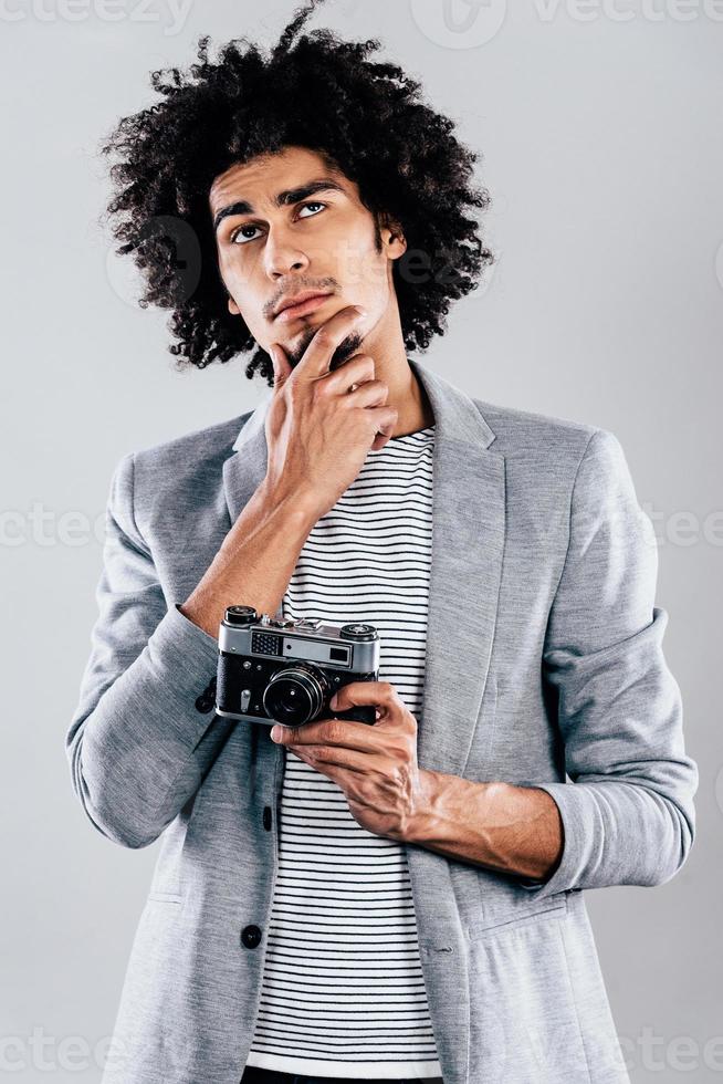 que dois-je tirer ensuite beau jeune homme africain tenant un appareil photo de style rétro et regardant pensif en se tenant debout sur fond gris