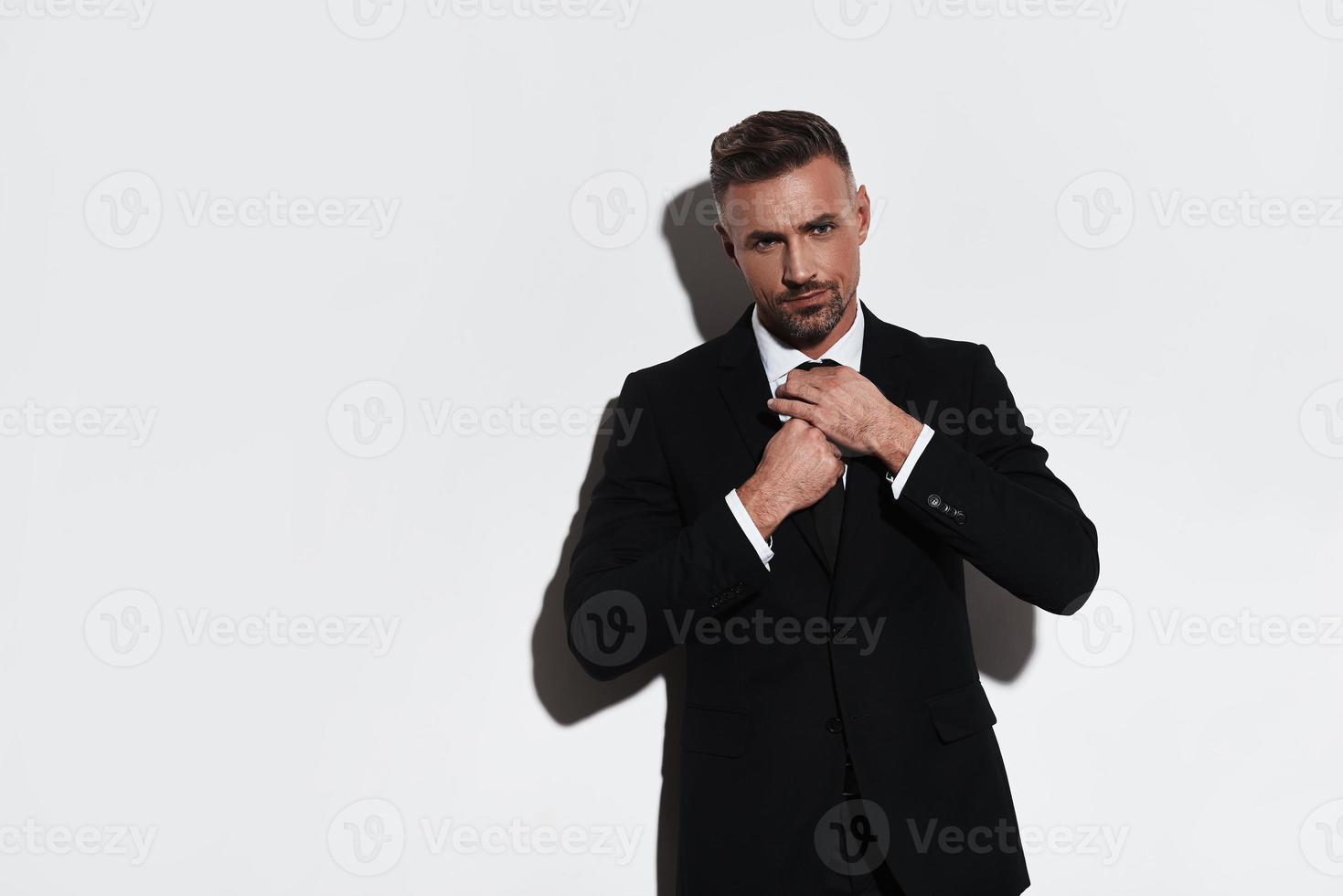 homme d'affaires confiant. beau jeune homme en costume complet ajustant la cravate et regardant la caméra en se tenant debout sur fond blanc photo