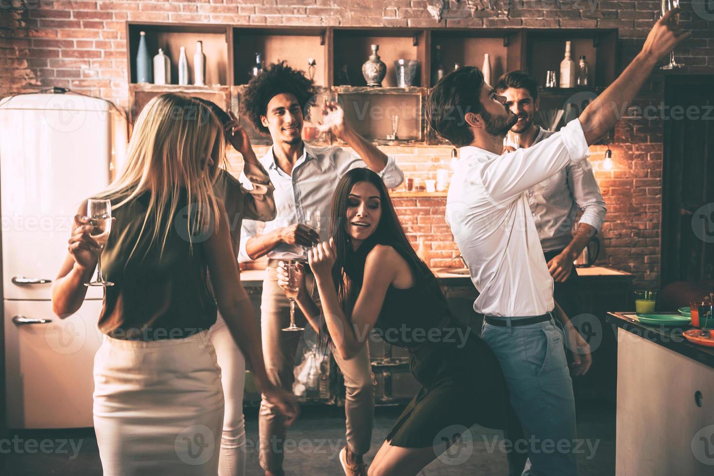 jouer dur. jeunes joyeux dansant et buvant tout en profitant d'une fête à la maison dans la cuisine photo