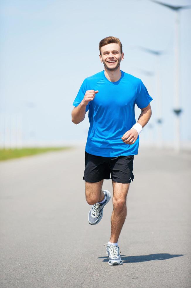 profiter de son jogging quotidien. vue de face pleine longueur du beau jeune homme qui court le long de la route et souriant photo