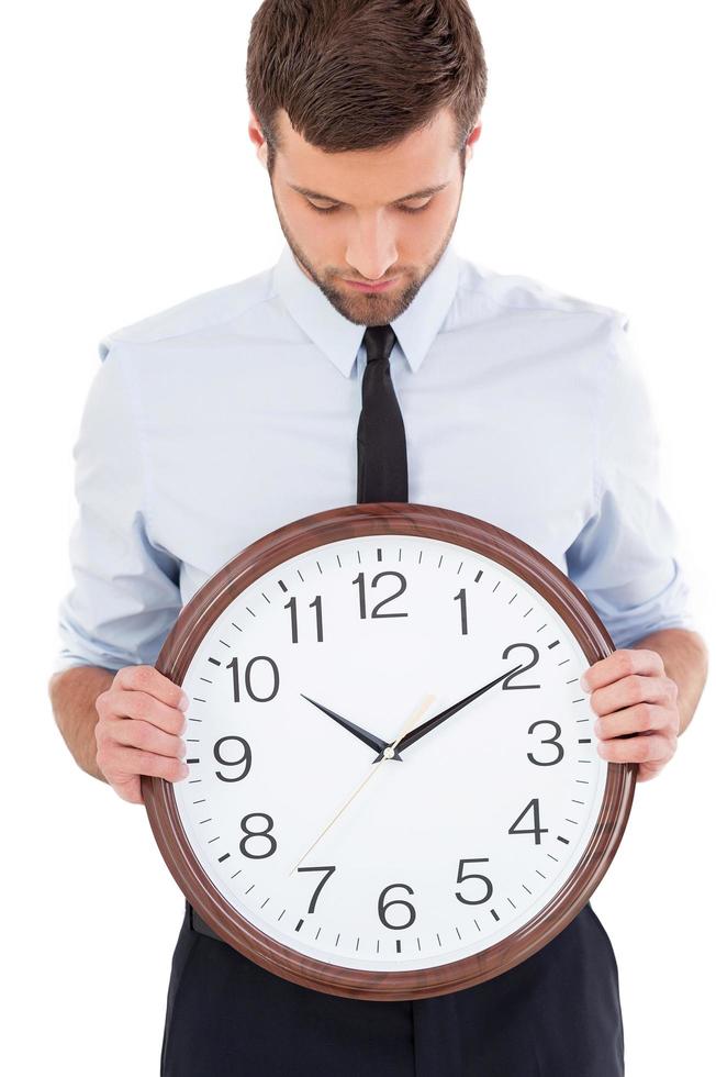 vérifier l'heure. Beau jeune homme en chemise et cravate tenant une horloge dans ses mains et en regardant tout debout isolé sur fond blanc photo