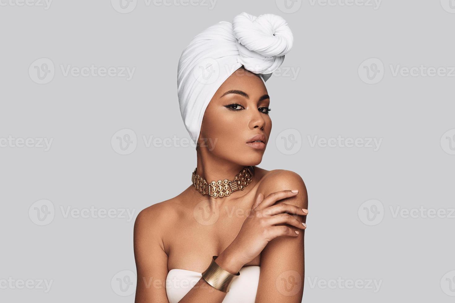 façon africaine. séduisante jeune femme africaine en turban regardant la caméra en se tenant debout sur fond gris photo