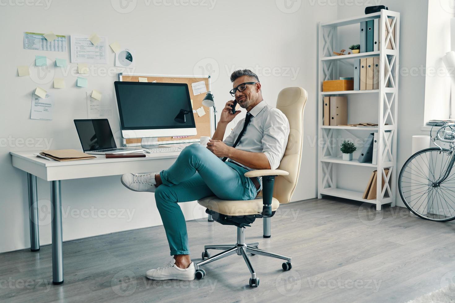 beau jeune homme en chemise et cravate parlant au téléphone intelligent et souriant assis au bureau photo