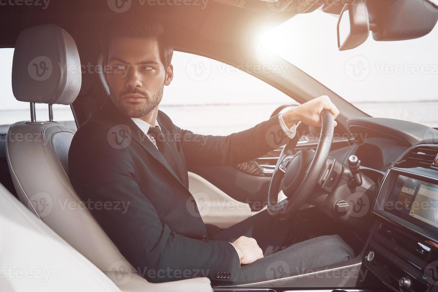 conducteur occupé. beau jeune homme en costume complet regardant par-dessus l'épaule en conduisant une voiture photo
