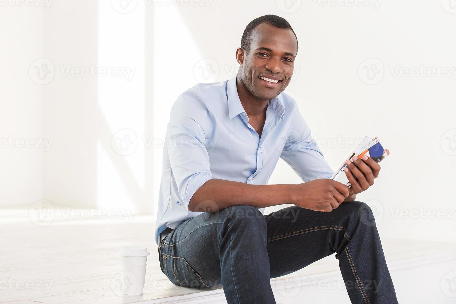 avoir une pause-café. joyeux jeune homme africain en chemise bleue tenant un magazine assis sur le sol avec une tasse de café près de lui photo
