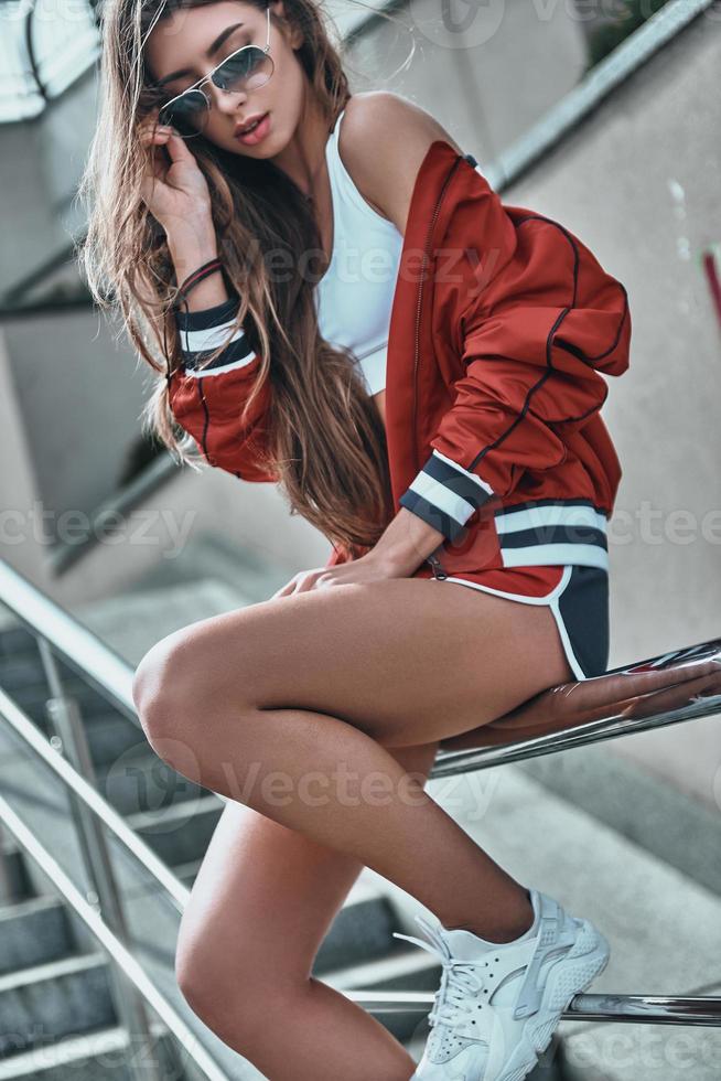 allure sportive. jolie jeune femme en vêtements de sport ajustant ses lunettes de soleil tout en étant assise sur la balustrade à l'extérieur photo