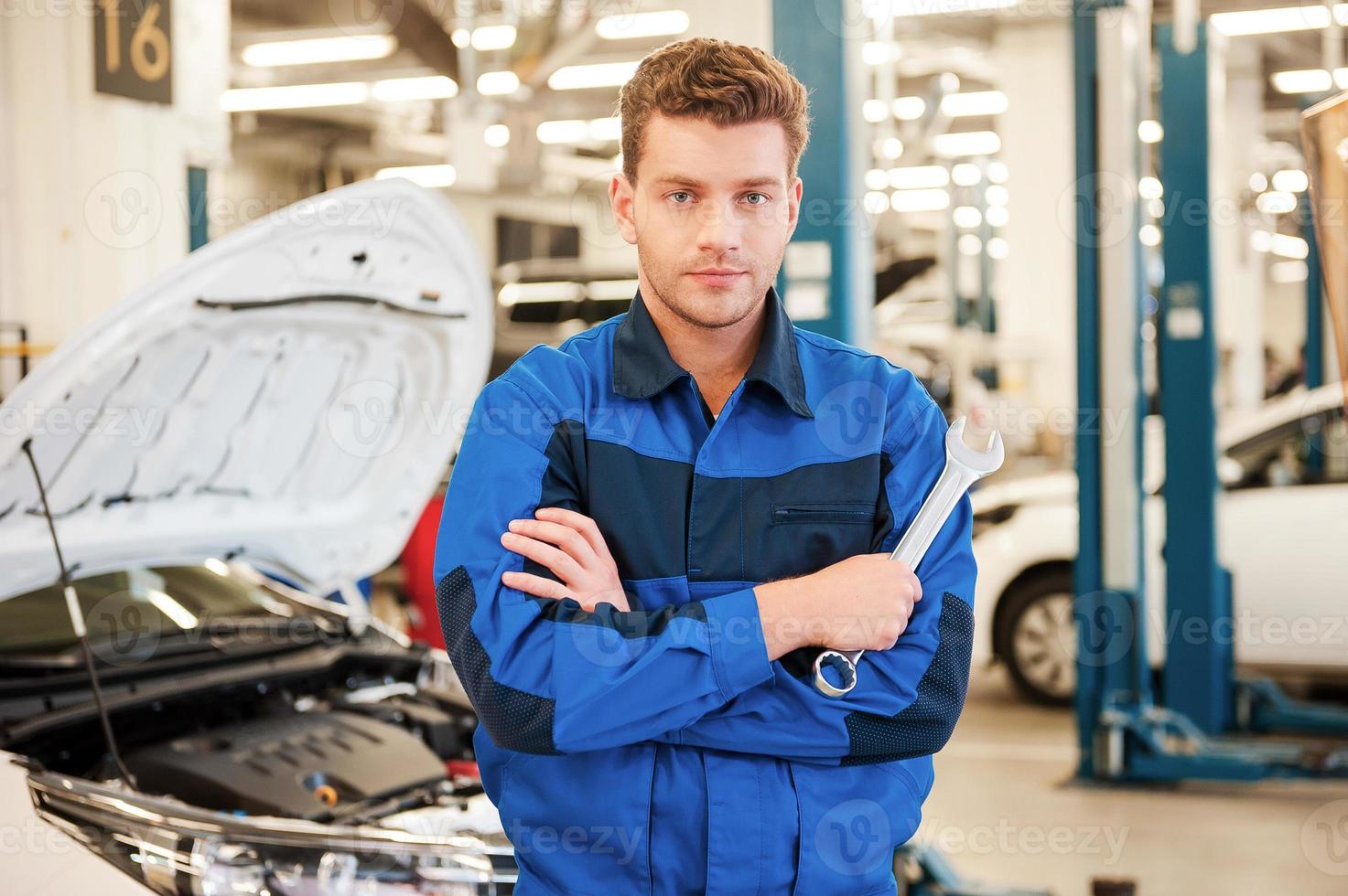 spécialiste confiant. jeune homme confiant tenant une clé et regardant la caméra tout en se tenant dans l'atelier avec la voiture en arrière-plan photo