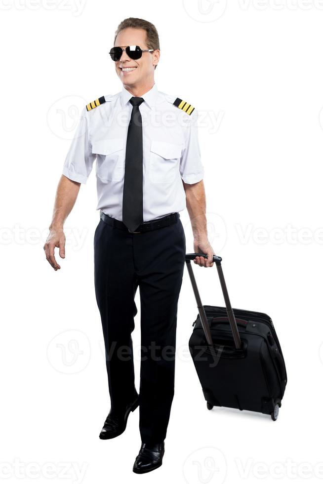 pilote joyeux. pilote masculin confiant en uniforme marchant et portant une valise tout en étant isolé sur fond blanc photo