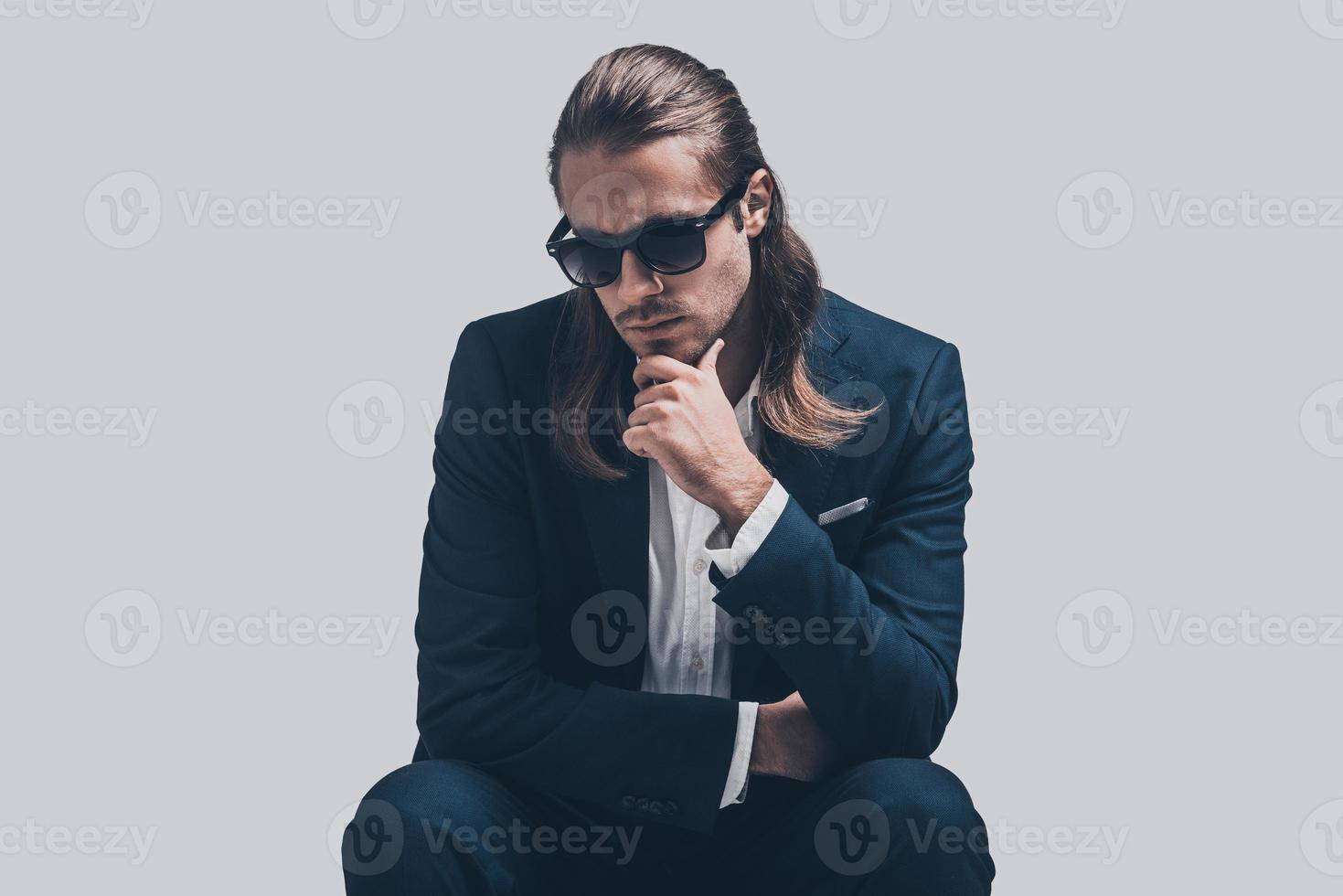 élégant et beau. beau jeune homme en costume complet et lunettes de soleil tenant la main sur le menton et l'air pensif assis sur fond gris photo