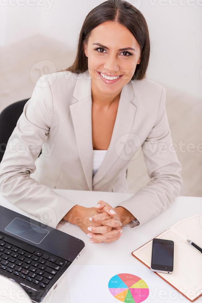 femme d'affaires heureuse. vue de dessus de la belle jeune femme d'affaires tenant les mains jointes et souriant alors qu'il était assis sur son lieu de travail photo