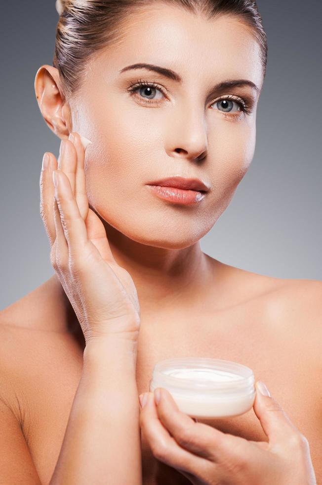 rester jeune et frais. portrait d'une femme mûre torse nu répandant de la crème sur le visage et regardant la caméra en se tenant debout sur fond gris photo