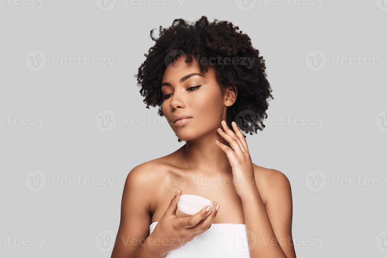 propre et frais. séduisante jeune femme africaine appliquant une crème hydratante et souriant en se tenant debout sur fond gris photo