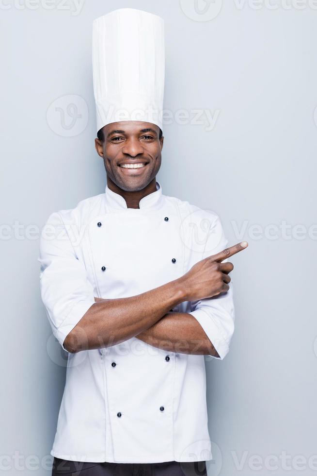 offre spéciale du chef. confiant jeune chef africain en uniforme blanc pointant vers l'extérieur et souriant en se tenant debout sur fond gris photo