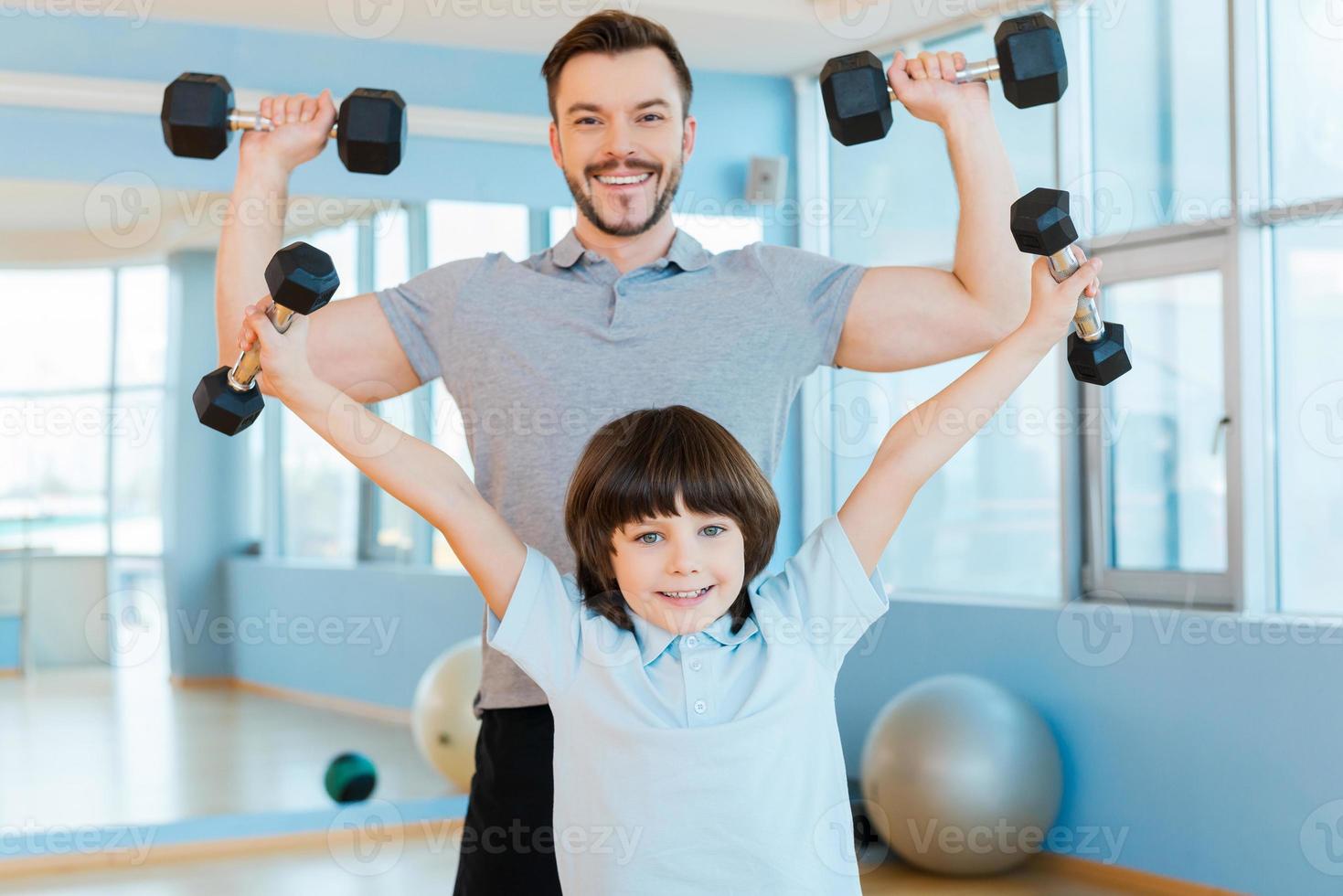 aussi fort que mon père. heureux père et fils faisant de l'exercice avec des haltères et regardant la caméra tout en se tenant tous les deux dans un club de santé photo