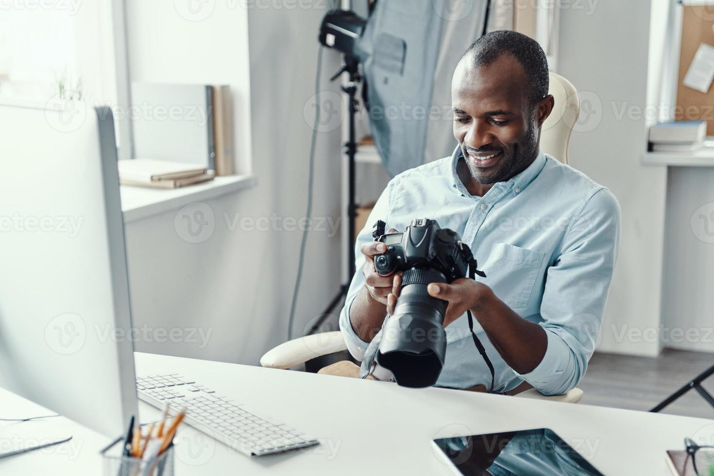 beau jeune homme africain tenant un appareil photo numérique et souriant tout en travaillant dans le bureau moderne