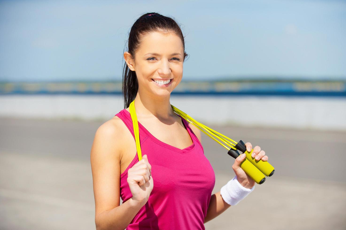 beauté avec corde à sauter. toute la longueur de la belle jeune femme en vêtements de sport tenant la corde à sauter et souriant tout en se tenant à l'extérieur photo