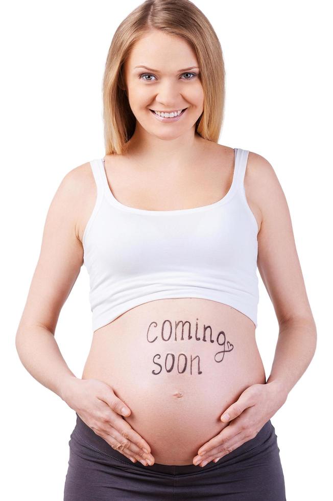 Bientôt disponible. image recadrée de femme enceinte avec bientôt signe sur son ventre debout isolé sur blanc photo