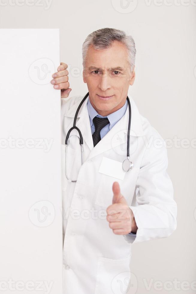 être en bonne santé médecin senior cheveux gris en uniforme regardant par l'espace de copie et faisant des gestes tout en étant isolé sur blanc photo