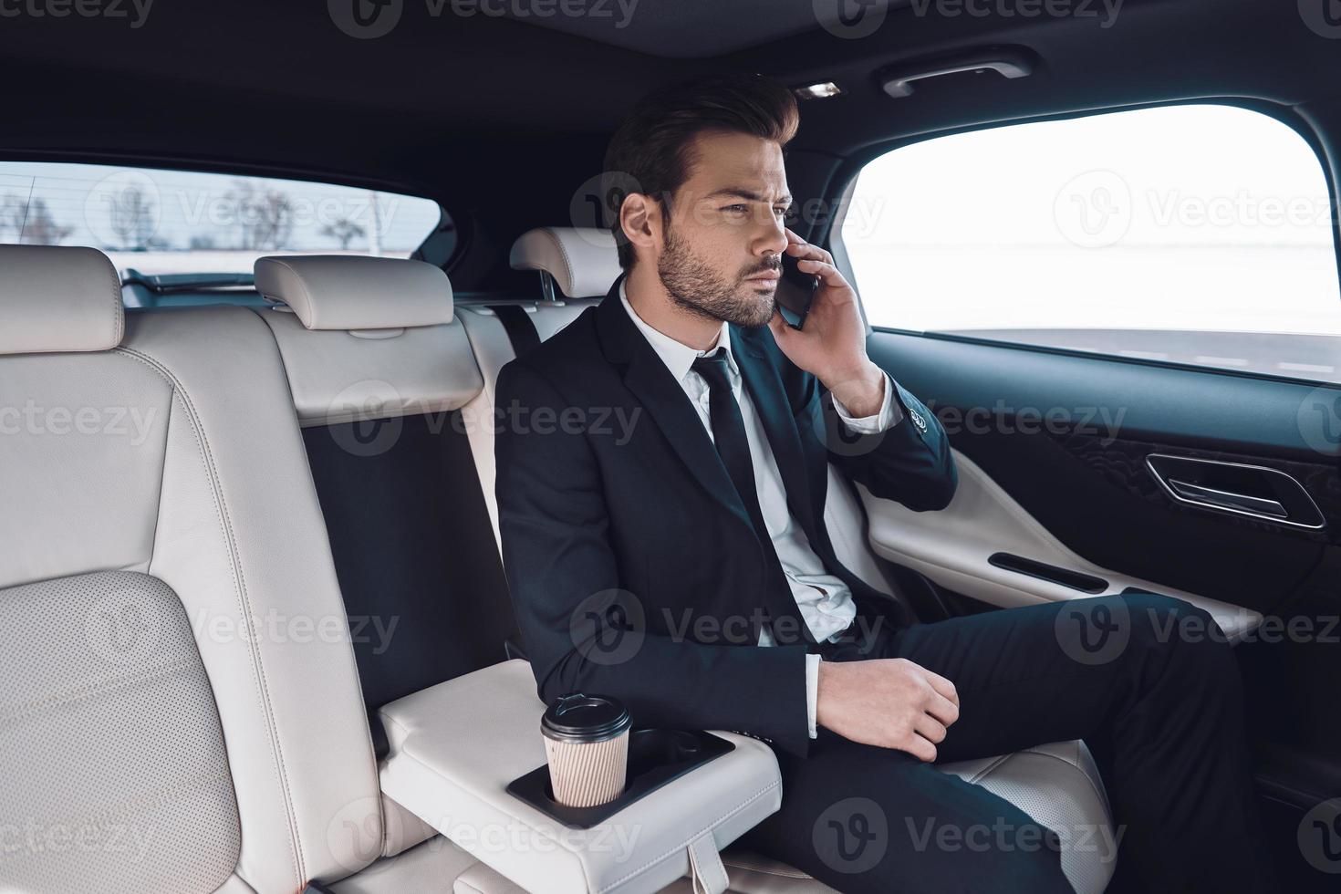 entreprise en mouvement. beau jeune homme en costume complet parlant au téléphone intelligent alors qu'il était assis dans la voiture photo