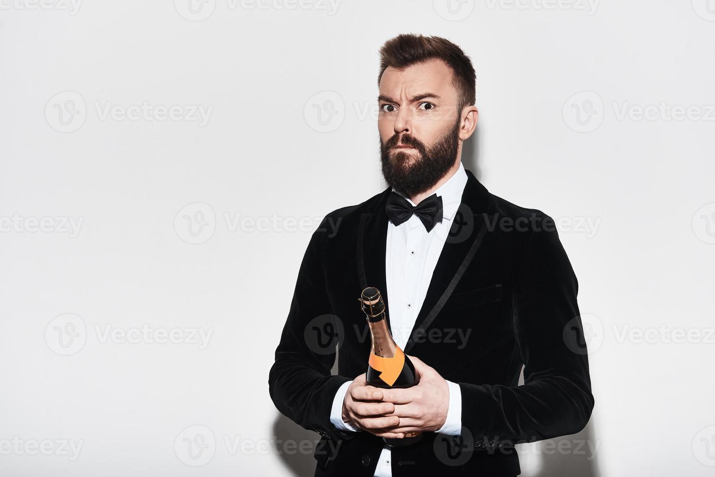 a quelque chose en tête. surpris jeune homme en costume complet portant une bouteille de champagne en se tenant debout sur fond gris photo