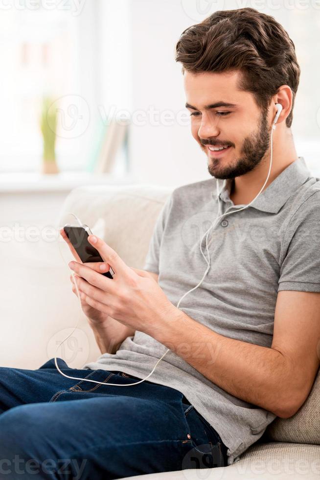 détente à la maison. heureux jeune homme barbu dans un casque tenant un lecteur mp3 et le regardant tout en étant assis sur un canapé photo