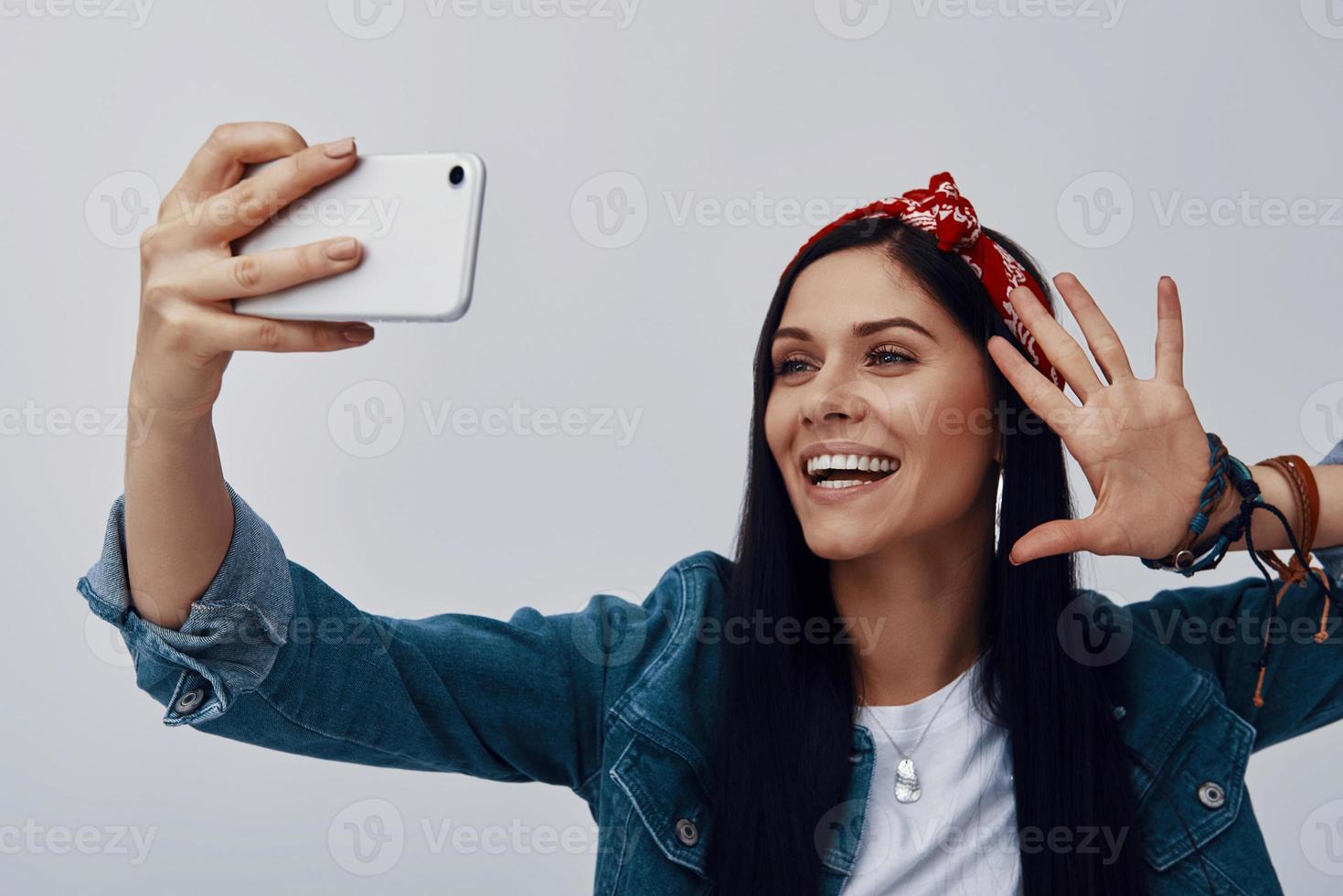 funky jeune femme en bandana faisant selfie et souriant debout sur fond gris photo