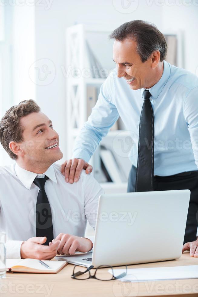partager une bonne expérience. homme d'affaires joyeux assis sur son lieu de travail pendant que son chef touche son épaule et se tient près de photo