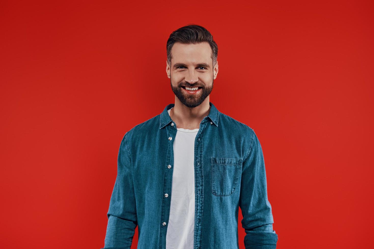 beau jeune homme en vêtements décontractés souriant et regardant la caméra en se tenant debout sur fond rouge photo