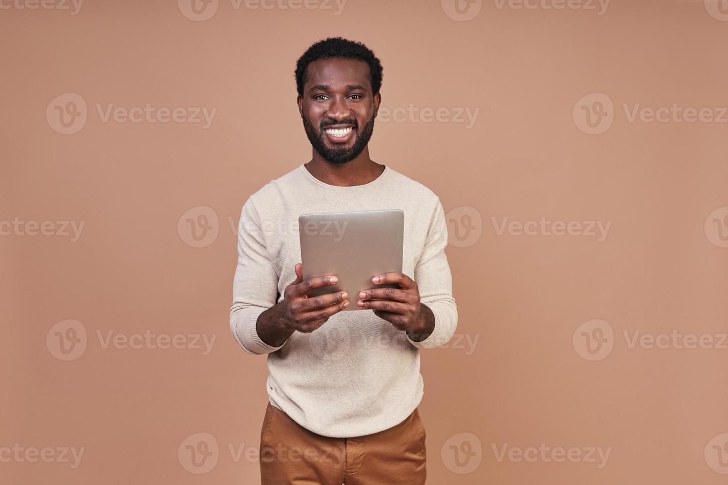 beau jeune homme africain en vêtements décontractés à l'aide d'une tablette numérique et souriant photo