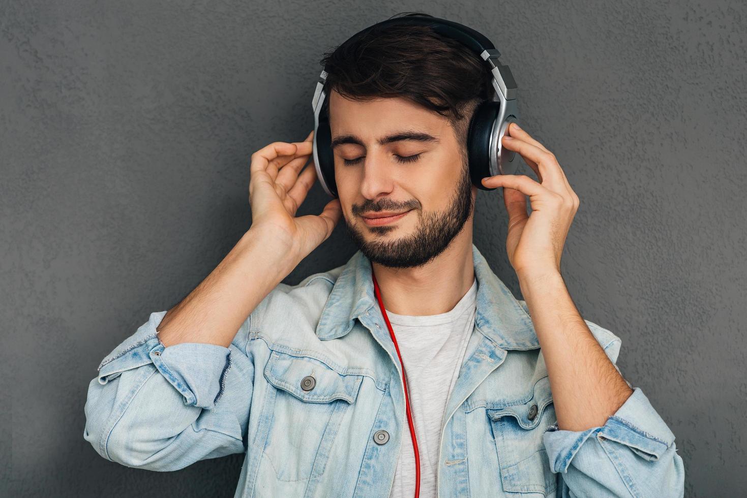 sa chanson préférée. jeune homme ajustant ses écouteurs et gardant les yeux fermés en se tenant debout sur fond gris photo