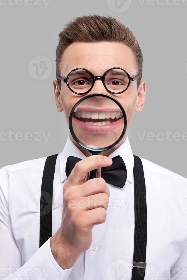 sourire grossissant. portrait d'un jeune homme joyeux en noeud papillon et bretelles tenant une loupe devant sa bouche et souriant photo