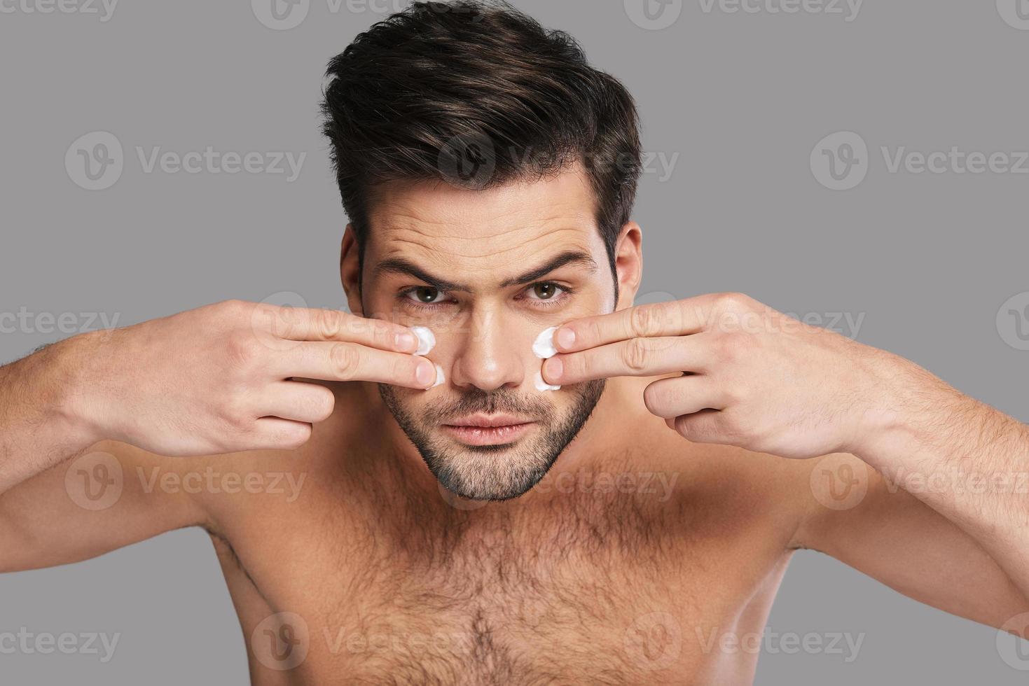 en prenant bien soin de sa peau. beau jeune homme appliquant une crème hydratante et regardant la caméra en se tenant debout sur fond gris photo