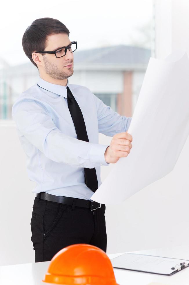 architecte au travail. jeune homme confiant en chemise et cravate examinant le plan tout en se tenant près de la table avec un casque dessus photo