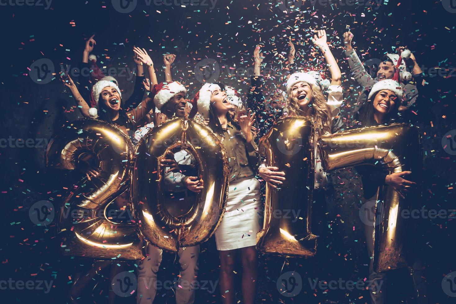 la nouvelle année 2017 arrive groupe de jeunes joyeux en chapeaux de père noël portant des numéros de couleur or et jetant des confettis photo