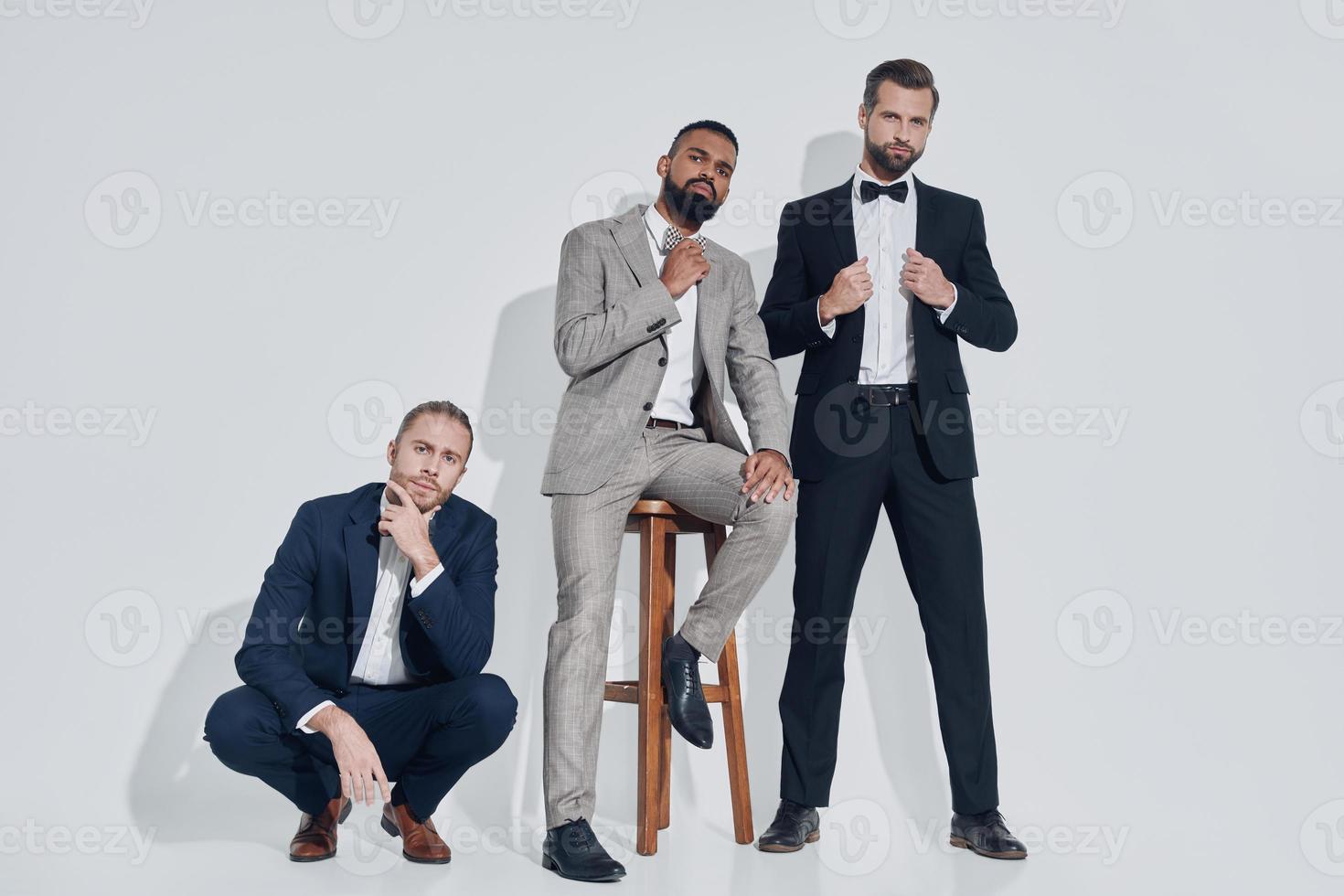 trois beaux jeunes hommes en costumes et noeuds papillon regardant la caméra en se tenant debout sur fond gris photo