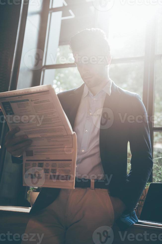 lire un journal frais. vue en angle bas d'un jeune homme confiant lisant un journal frais tout en se penchant sur le rebord de la fenêtre au bureau ou au café photo