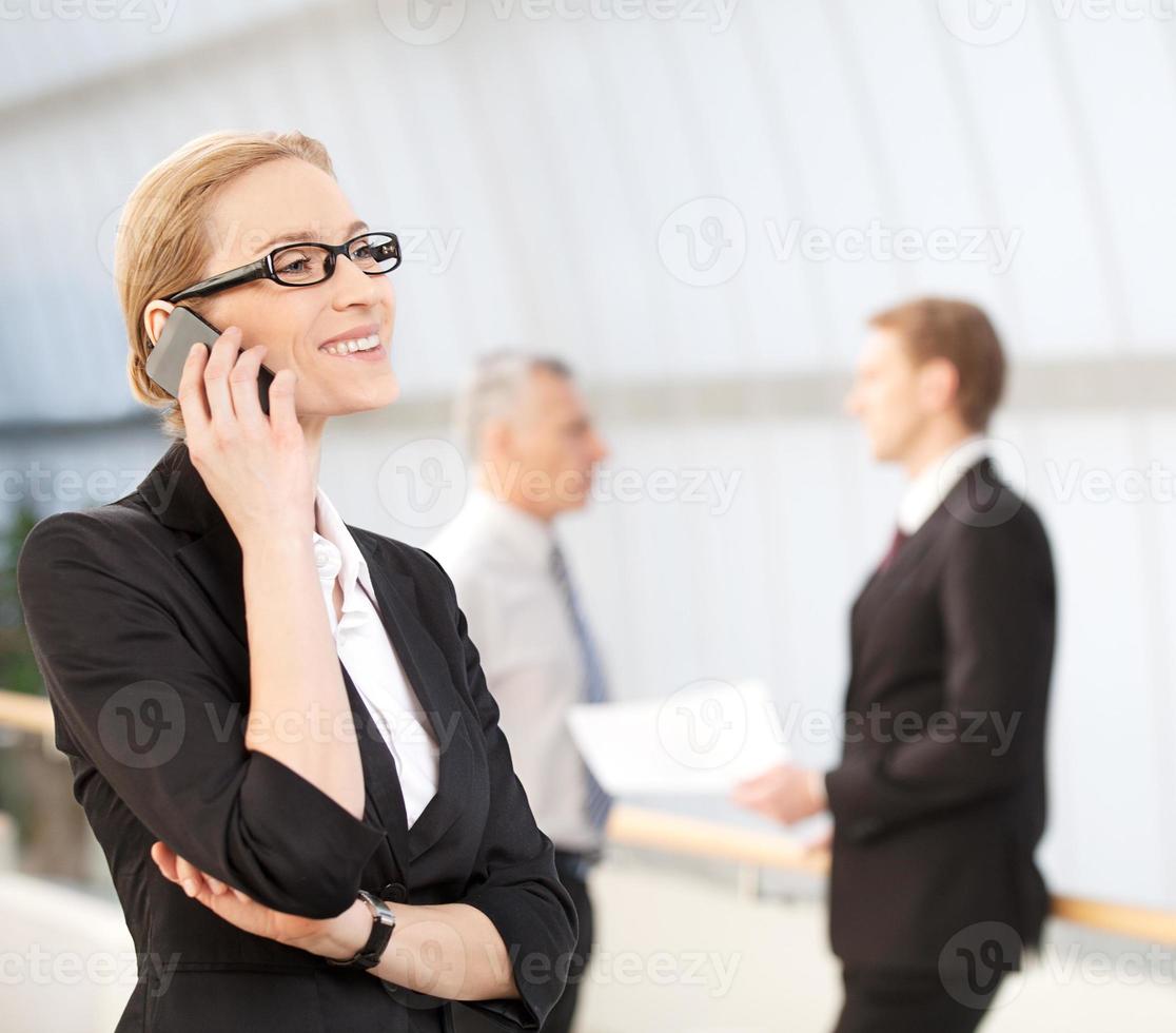 femme d'affaires au téléphone. femme mûre joyeuse en tenues de soirée parlant au téléphone mobile et souriant pendant que ses collègues parlent en arrière-plan photo