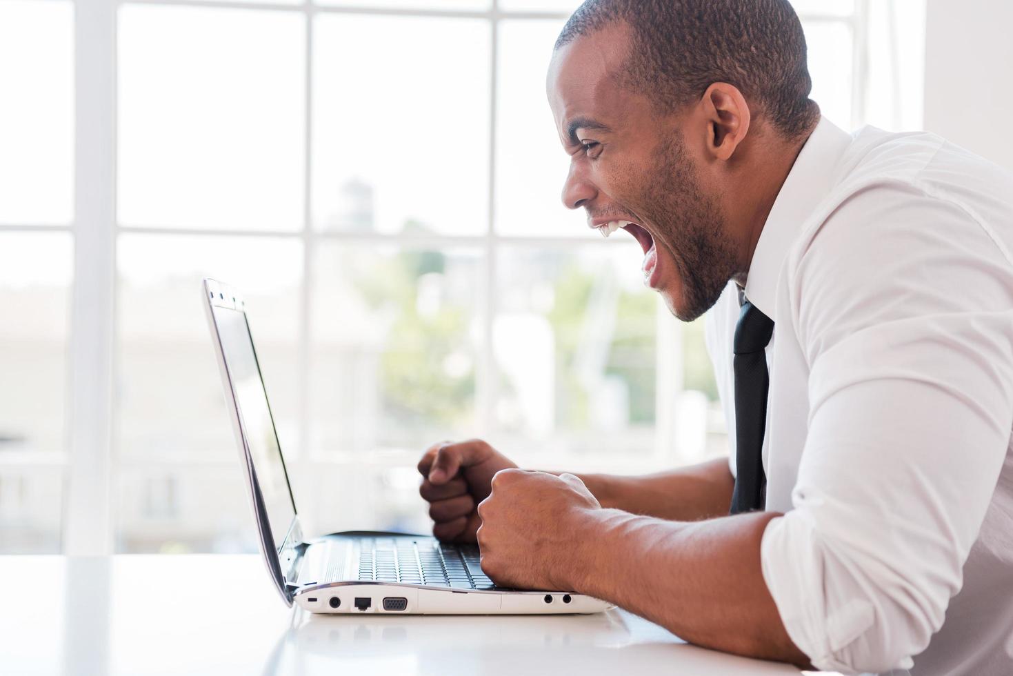 employé de bureau stressé. vue latérale d'un jeune homme africain furieux en chemise et cravate criant tout en regardant un ordinateur portable assis sur son lieu de travail photo