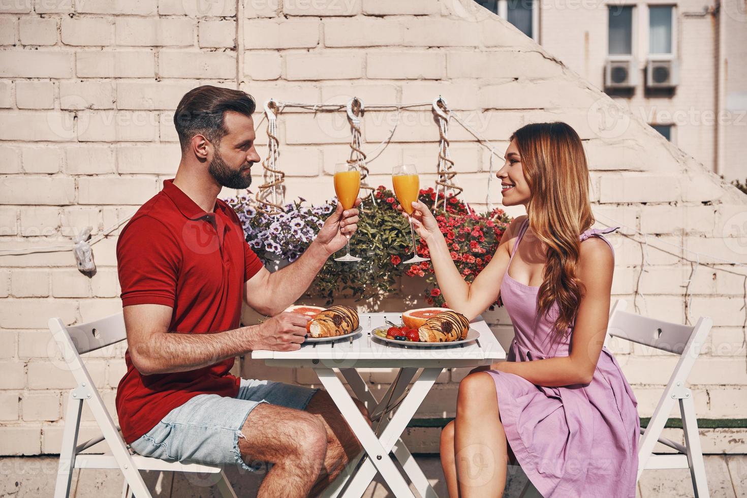 jeune couple moderne en vêtements décontractés portant un toast et souriant tout en étant assis sur la terrasse sur le toit à l'extérieur photo