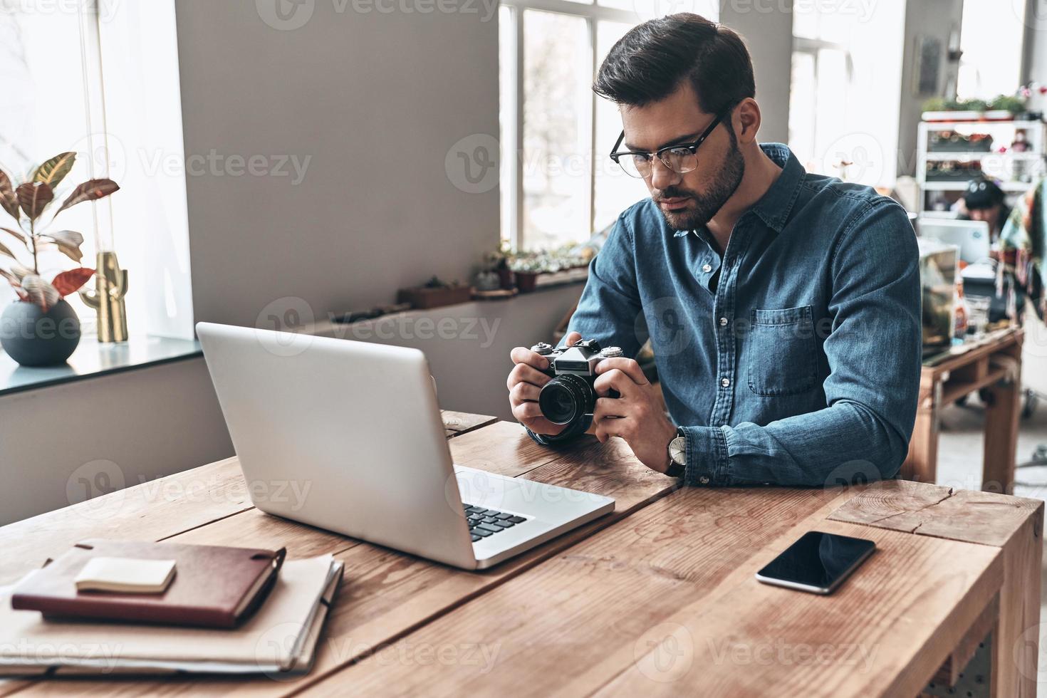 jeune perfectionniste. jeune homme réfléchi utilisant un appareil photo tout en travaillant dans l'espace de travail créatif