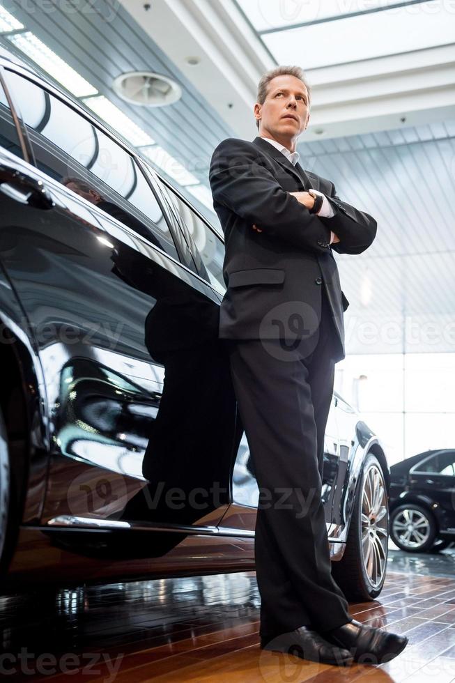 vivre une vie de luxe. vue en angle bas d'un homme confiant aux cheveux gris en tenue de soirée se penchant sur la voiture et regardant au loin photo