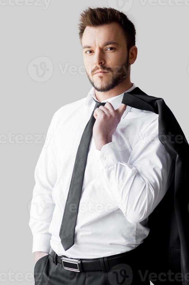 homme d'affaires détendu. beau jeune homme en tenues de soirée tenant sa veste sur le doigt et regardant la caméra en se tenant debout sur fond gris photo